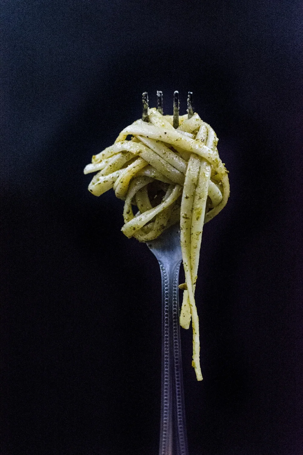 A fork holding a serving of spaghetti with a green seasoning, against a black background.