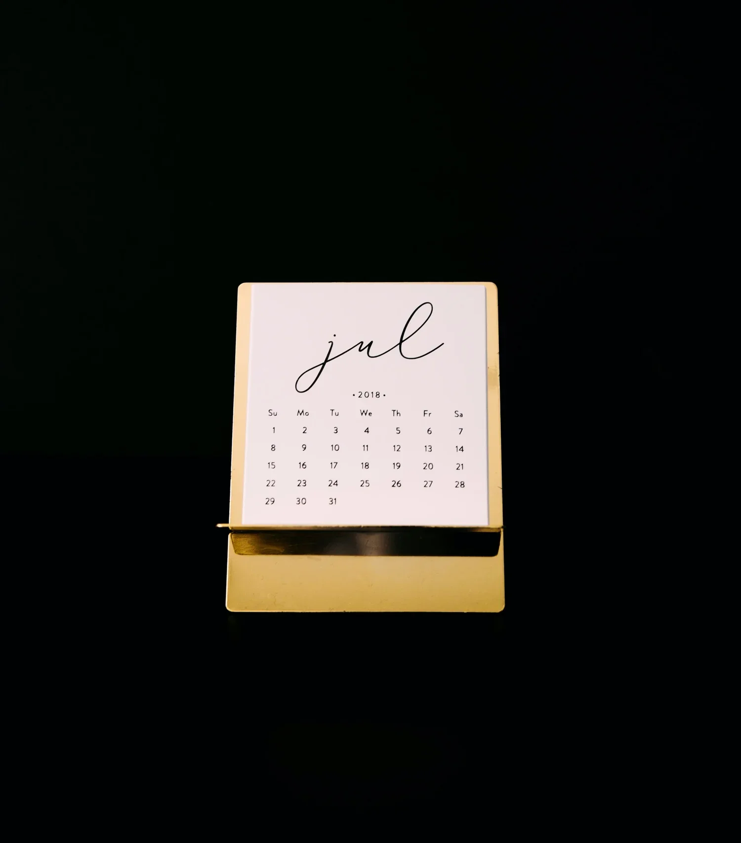 A desk calendar for July 2018 with a white background and black cursive text, placed on a reflective surface against a black background.
