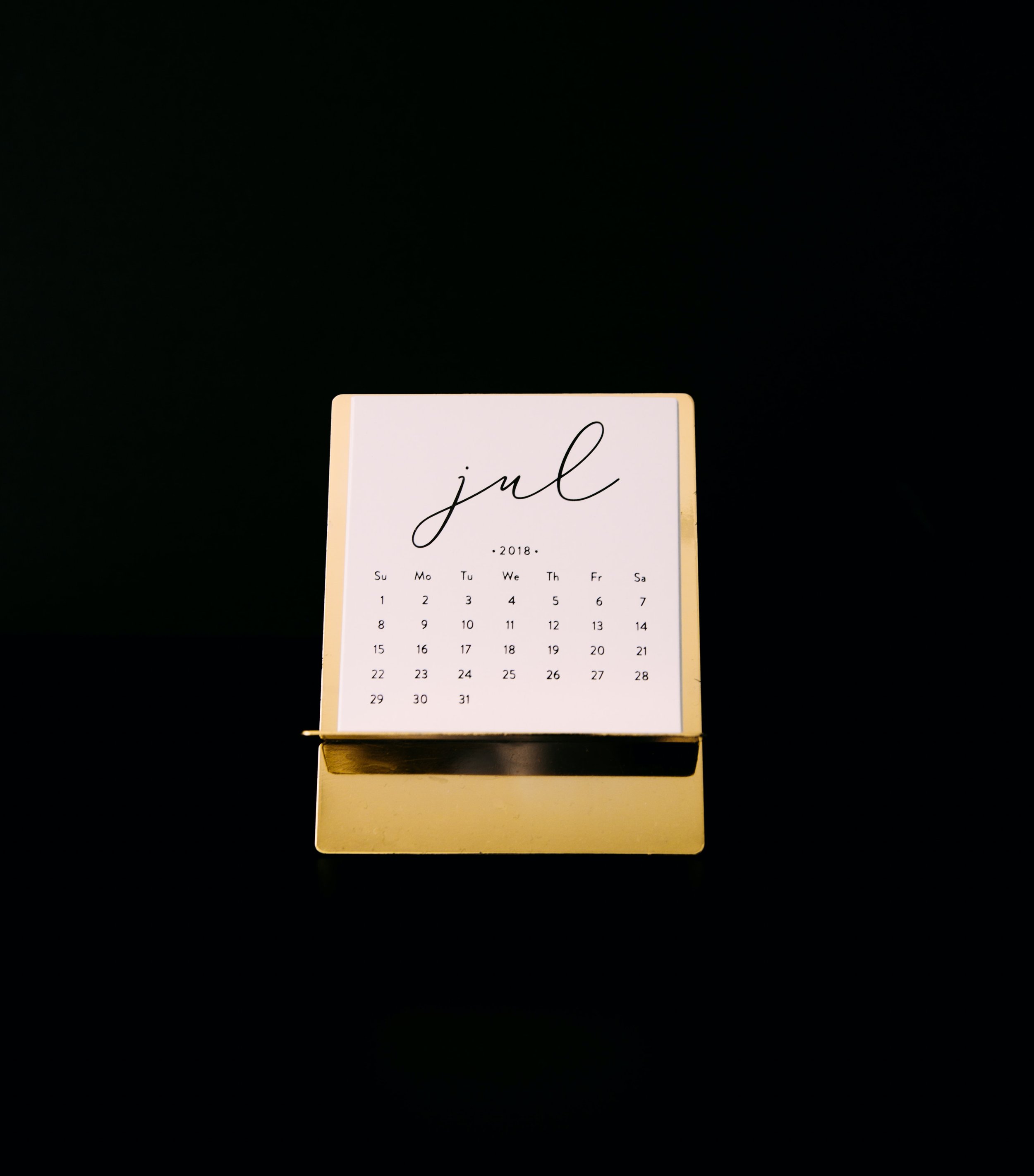 A small desk calendar displays the month of July 2018, with the days of the week labeled in abbreviated form from Sunday to Saturday, set against a black background.