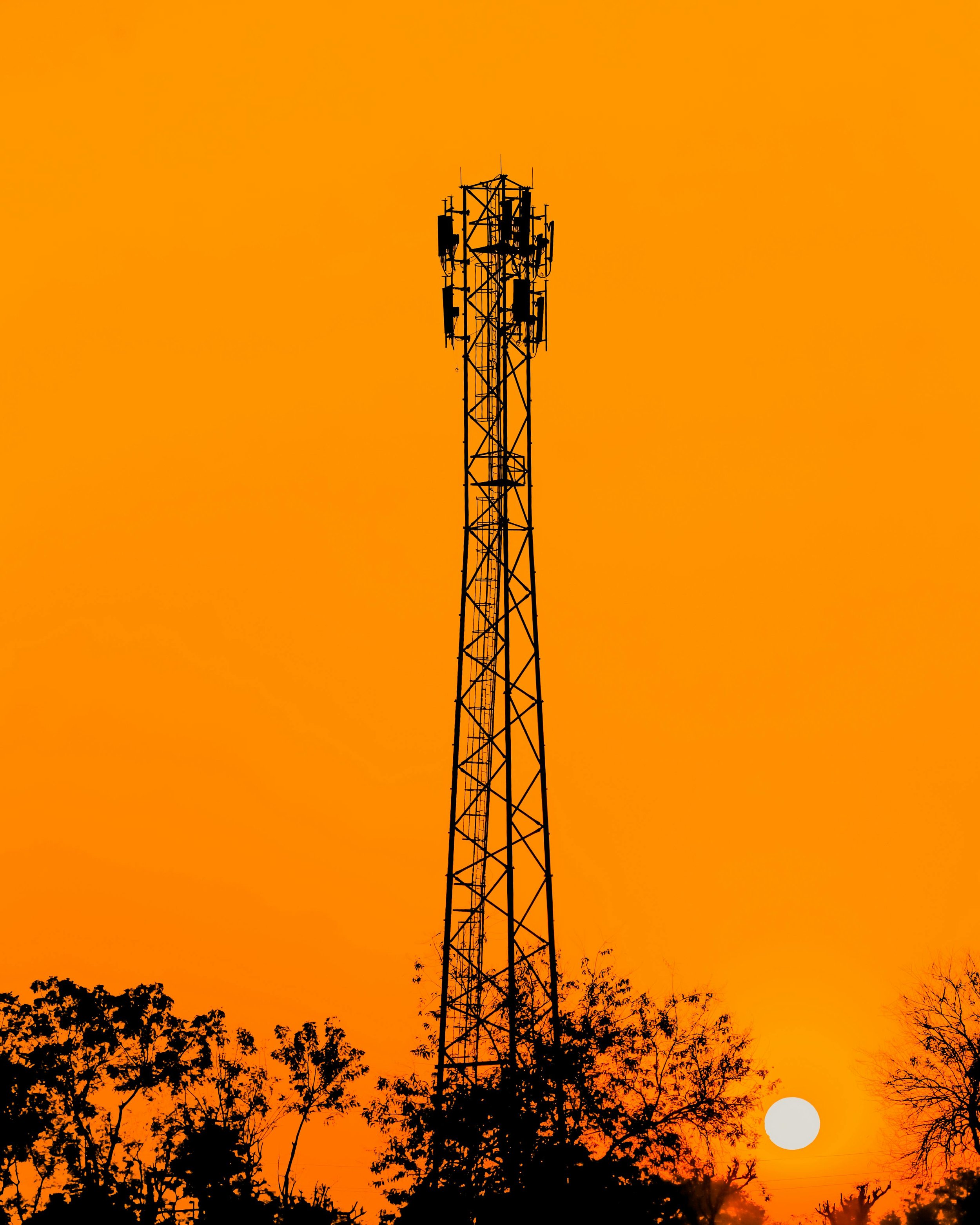 Silhouette of a cell tower with antennas and equipment against an orange sunset sky, with trees in the foreground and a white sun near the bottom right corner.