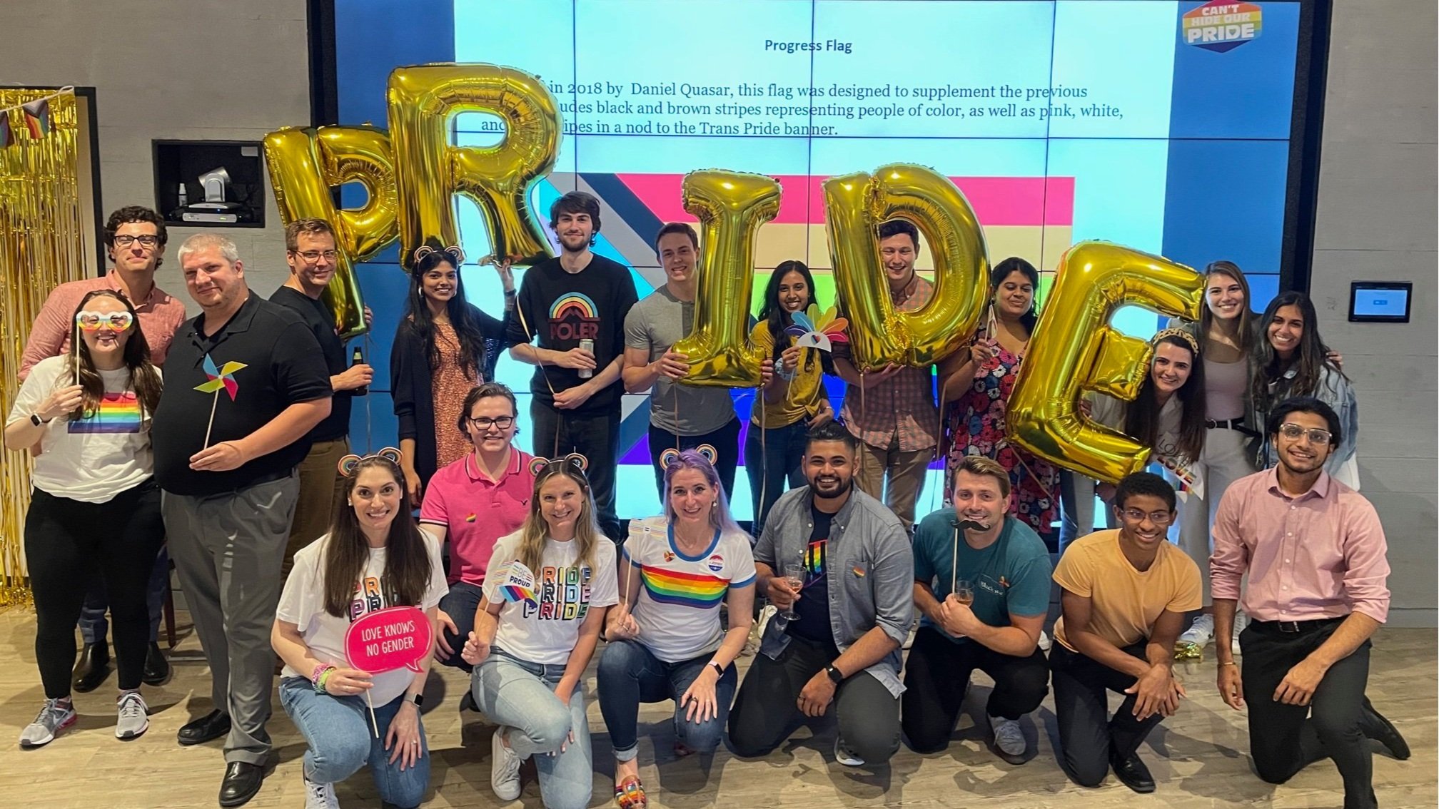 A group of people celebrating Pride, holding rainbow flags, and balloons spelling out 'PRIDE'. Some individuals wear pride-themed clothing and accessories, and there is a large digital screen in the background with Pride-related information and icons.